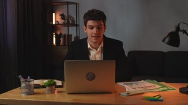 Günlük rutin yaşam tarzı konsepti. Ofisteki genç işadamı geç saatlere kadar çalışıyor. Masada oturan, bilgisayarla konuşan, bilgisayarla konuşan iş arkadaşlarını selamlıyor..