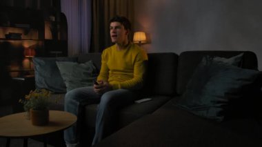 Günlük rutin yaşam tarzı konsepti. Akşamları işten sonra evde dinlenen, oturma odasındaki koltukta oturan uzaktan kumandayla televizyon kanallarını değiştiren, spor maçı izleyen, tezahürat yapan ve konuşan genç bir adam..