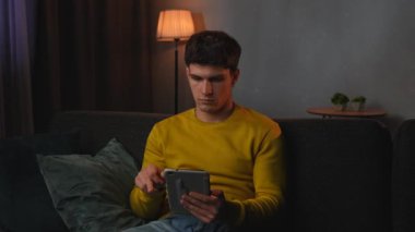 Günlük rutin yaşam tarzı konsepti. Akşamları işten sonra evde dinlenen, oturma odasında kanepede oturup televizyon izleyen, elinde tabletle sosyal medyayı soyan genç bir adam..