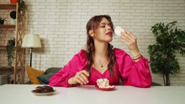 Tatlılar ve tatlılar reklam konsepti. Genç bir kadın masada oturuyor ve bisküvi yiyor, bir ısırık alıp çiğniyor, tatlı tatlı atıştırmanın tadını çıkarıyor, mutlu bir ifade..