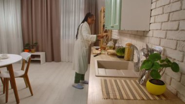 Günlük rutin yaşam tarzı konsepti. Genç Afro-Amerikalı kadın sabah mutfakta sağlıklı bir kahvaltı hazırlıyor. Ekmeğe böğürtlen reçeli sürüyor..