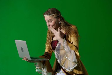 Tarihsel reklam konsepti. Krom anahtar yeşil ekran arka planda antik kıyafetli bir kadın. Rönesans elbisesi giymiş bir kadın. Şaşırmış bir yüz ve elinde dizüstü bilgisayarla ekrana bakıyor..