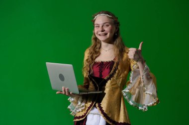 Tarihsel reklam konsepti. Krom anahtar yeşil ekran arka planda antik kıyafetli bir kadın. Rönesans elbisesi giymiş, mutlu yüzlü bir kadın başparmağını kaldırmış, elinde dizüstü bilgisayarla kameraya bakıyor..