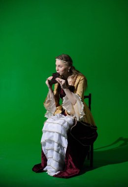 Tarihsel reklam konsepti. Krom anahtar yeşil ekran arka planda antik kıyafetli bir kadın. Rönesans elbisesi giymiş bir kadın oturmuş, büyük çikolatalar yiyor, ısırıyor ve eğleniyor..