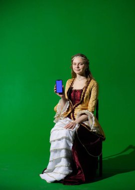 Tarihsel reklam konsepti. Krom anahtar yeşil ekran arka planda antik kıyafetli bir kadın. Rönesans elbisesi giymiş bir kadın elinde mavi reklam ekranlı akıllı bir telefonla oturuyor..