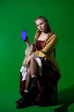 Tarihsel reklam konsepti. Krom anahtar yeşil ekran arka planda antik kıyafetli bir kadın. Rönesans elbisesi giymiş bir kadın elinde mavi reklam ekranlı akıllı bir telefonla oturuyor..