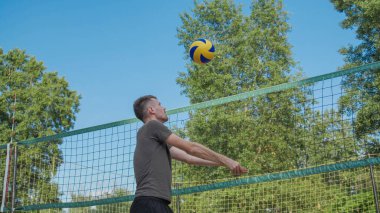 Gri tişörtlü ve siyah şortlu genç bir adam voleybol sahasında voleybol oynuyor. Bir yaz öğleden sonrasında, mavi gökyüzünün altındaki yeşil ağaçlarla çevrili olarak ağın başında duruyor.