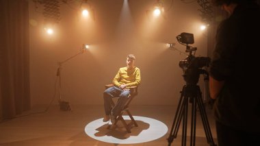 Profesyonel bir stüdyonun içinde. Aydınlatma ekipmanları ve siklonamayla fotoğraf çekimi stüdyosu, model aktör ortada yönetmen koltuğunda oturuyor ve röportaj kaydediliyor..