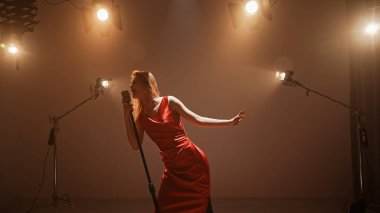 Profesyonel bir stüdyonun içinde. Işıklandırma ekipmanları ve siklonamayla fotoğraf çekimi stüdyosu. Ortadaki retro mikrofonda şarkı söyleyen kırmızı elbiseli kadın şarkıcı..