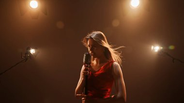 Profesyonel bir stüdyonun içinde. Işıklandırma ekipmanları ve siklonamayla fotoğraf çekimi stüdyosu. Ortadaki retro mikrofonda şarkı söyleyen kırmızı elbiseli kadın şarkıcı..