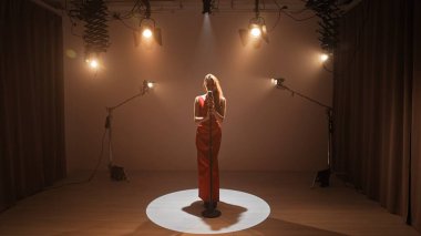 Profesyonel bir stüdyonun içinde. Işıklandırma ekipmanları ve siklonamayla fotoğraf çekimi stüdyosu. Ortadaki retro mikrofonda şarkı söyleyen kırmızı elbiseli kadın şarkıcı..