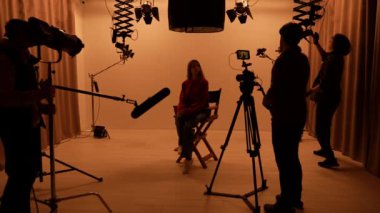 Profesyonel stüdyonun içi. Işık ekipmanları ve siklonamayla fotoğraf çekimi stüdyoları, ışık ve kamera hazırlayan ekip üyeleri, ortada oturan yönetmen koltuğunda oturan mankenler..