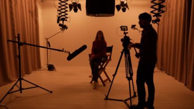 Profesyonel bir stüdyonun içinde. Işık ekipmanları ve siklonamayla fotoğraf çekimi stüdyosu, ışık ve kamera hazırlayan ekip üyeleri, model yönetmen koltuğunda oturup röportaj yapıyor..