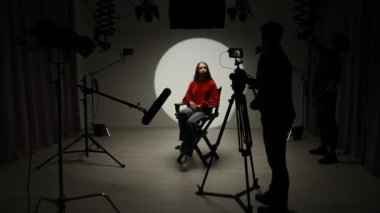 Profesyonel bir stüdyonun içinde. Işık ekipmanları ve siklonamayla fotoğraf çekimi stüdyosu, ışık ve kamera hazırlayan ekip üyeleri, model yönetmen koltuğunda oturup röportaj yapıyor..