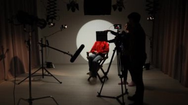Profesyonel bir stüdyonun içinde. Aydınlatma ekipmanları ve siklonamayla fotoğraf çekimi stüdyosu, model aktris ortada yönetmen koltuğunda oturuyor ve röportajı kaydediyor..