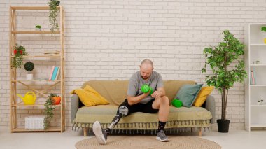 Protez bacaklı bir adam rahat bir oturma odasında kanepede oturur, dambıllarla egzersiz yapar, kollarını çalıştırır. Kısıtlı hareketli adaptif yaşam kavramı.