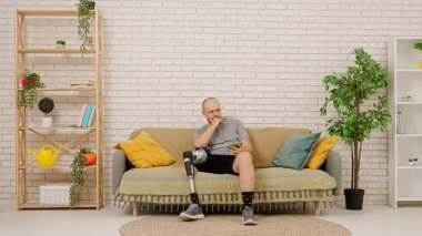 Protez bacaklı bir adam rahat bir oturma odasında kanepede oturup akıllı telefonuyla video izliyor ve kaybettiği için üzülüyor. Kısıtlı hareketli adaptif yaşam kavramı.