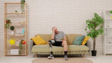 Protez bacaklı bir adam rahat bir oturma odasında kanepede oturup akıllı telefonuyla video izliyor ve kaybettiği için üzülüyor. Kısıtlı hareketli adaptif yaşam kavramı.