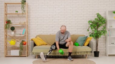 Protez bacaklı bir adam rahat bir oturma odasında kanepede oturur, dambıllarla egzersiz yapar, kollarını çalıştırır. Kısıtlı hareketli adaptif yaşam kavramı.