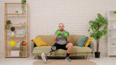 Protez bacaklı bir adam rahat bir oturma odasında kanepede oturur, dambıllarla egzersiz yapar, kollarını çalıştırır. Kısıtlı hareketli adaptif yaşam kavramı.