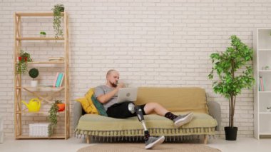 Oturma odasındaki kanepede oturan protez bacaklı bir adam dizüstü bilgisayardaki bilgilere bakıyor ve bir deftere yazı yazıyor. Kısıtlı hareketli adaptif yaşam biçimi kavramı.