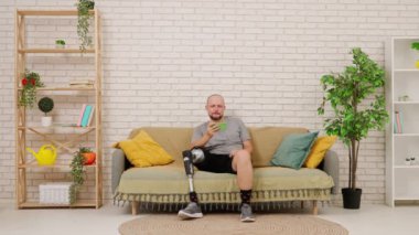 Protez bacaklı bir adam rahat bir oturma odasında kanepede oturur, akıllı telefonuyla video izler ve zaferle sevinir. Kısıtlı hareketli adaptif yaşam kavramı.