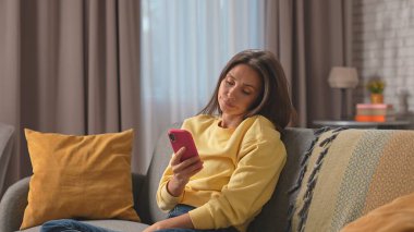 Kadın evde boş vakit geçiriyor. Genç bir kadın, oturma odasındaki kanepede oturmuş elinde akıllı bir telefonla sosyal medyayı karıştırıyor, ilgisiz bir ifadeyle sıkılıyor..