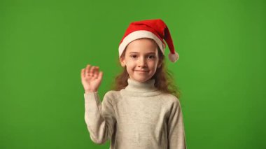 Çocuk duyguları konsepti. Noel Baba şapkalı gülümseyen kız kameraya ve el sallamasına, olumlu ifadeye, krom anahtar yeşil ekranlı arka plan modeline bakıyor..