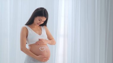 Genç hamile kadın cildine bakarken çıplak karnına krem sürüyor. Müstakbel anne stüdyoda beyaz perdelere karşı poz veriyor. Annelik kavramı.