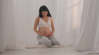 Asyalı genç hamile kadın elleriyle karnını okşuyor. Müstakbel anne stüdyoda beyaz perdelere karşı poz veriyor. Annelik kavramı.