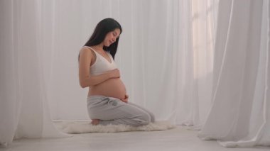 Genç hamile kadın elleriyle karnını nazikçe okşuyor. Müstakbel anne stüdyoda beyaz perdelere karşı poz veriyor. Annelik kavramı.