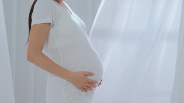 Beyaz elbiseli Asyalı hamile kadın elleriyle karnını okşuyor. Müstakbel anne stüdyoda beyaz perdelere karşı poz veriyor. Annelik kavramı.