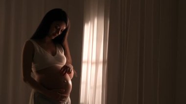 Asyalı genç hamile kadın elleriyle karnını okşuyor. Müstakbel anne stüdyoda akşam ışığında perdelere karşı poz veriyor. Annelik kavramı.