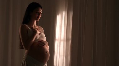 Doğumdan önce çıplak karnıyla nefes egzersizi yapan Asyalı hamile bir kadın. Müstakbel anne stüdyoda akşam ışığında perdelere karşı poz veriyor. Annelik kavramı.