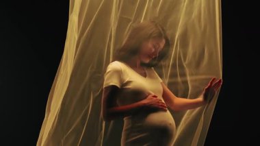 Beyaz elbiseli genç Asyalı hamile bir kadın şefkati ve anneliği simgeleyen akıcı bir kumaşın arkasında zarifçe poz veriyor. Müstakbel anne stüdyoda poz veriyor.