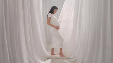 Beyaz elbiseli Asyalı hamile kadın elleriyle karnını okşuyor. Müstakbel anne stüdyoda beyaz perdelere karşı poz veriyor. Annelik kavramı.
