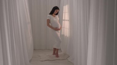 Beyaz elbiseli Asyalı hamile kadın elleriyle karnını okşuyor. Müstakbel anne stüdyoda beyaz perdelere karşı poz veriyor. Annelik kavramı.