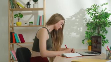 Çevrimiçi kursa katılan kişi. Evde çalışan bir üniversite öğrencisi, dizüstü bilgisayardan ders izliyor, not defterine yazıyor, masa başında oturuyor, fotoğraf çekiyor. Modern online eğitim.
