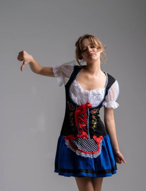 Genç bir kadın zarif bir şekilde korse, kabarık kollar ve dekoratif detaylarla süslenmiş mavi bir etekle Bavyera tarzı bir elbise içinde poz veriyor ve şenlikli bir halk atmosferini anımsatıyor. Baş parmak hareketi..