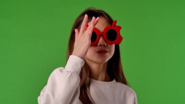 Çocuk duyguları konsepti. Asyalı kız çocuğu kameraya poz veriyor, kırmızı elma gözlüğü takıyor, pozitif ifade, krom anahtar yeşil ekran arka plan modelinde izole edilmiş..
