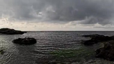 İspanya 'da kayalık bir kıyı şeridine yakın, berrak su kayaların üzerinden Akdeniz' e doğru yavaşça akıyor. Karanlık, ıslak kayalar yumuşak gökyüzü ile tezat oluşturarak sakin bir kıyı manzarası yaratıyor..