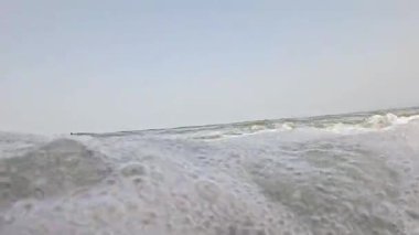 Güçlü bir okyanus dalgası kıyıya çarpıyor ve derin mavi suya karşı beyaz köpük oluşturuyor. Görüntü, açık bir gökyüzünün altında denizin ham enerjisini yakalıyor..