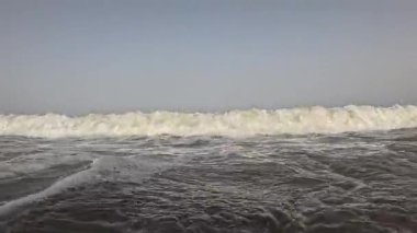 Güçlü bir okyanus dalgası kıyıya çarpıyor ve derin mavi suya karşı beyaz köpük oluşturuyor. Görüntü, açık bir gökyüzünün altında denizin ham enerjisini yakalıyor..