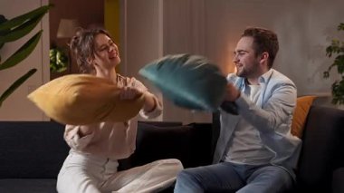 Mutlu çift birlikte vakit geçiriyor. Evdeki kanepede dinlenen sevimli bir çift yastık savaşında oynayan bir kadın ve bir erkek şakalaşıyor ve sarılıyor, olumlu ifade. Yavaş çekim.