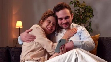 Mutlu çift birlikte vakit geçiriyor. Hoş bir çift evdeki kanepede dinleniyor, kadın ve erkek sarılıyor, akıllı telefondan sosyal medyaya kayıyor, mutlu kadın kazanan jest yapıyor ve erkeği öpüyor..