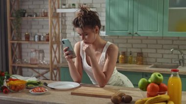 Sağlıklı beslenme konsepti. Mutfaktaki masada sağlıklı öğle yemeği için börek hazırlayan genç bir kadın akıllı telefondan mesaj atan, düşünen ve gülümseyen bir kadın. Yavaş çekim.