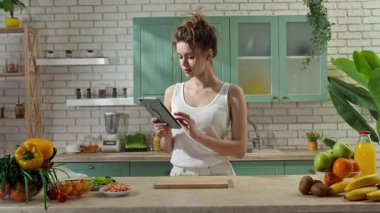 Sağlıklı beslenme konsepti. Mutfaktaki masada genç bir kadın sağlıklı öğle yemeği için börek hazırlıyor, tabletten yemek tarifi okuyor, abur cubur hazırlamak için aldığı malzemeleri kontrol ediyor. Yavaş çekim.