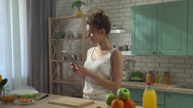 Sağlıklı beslenme konsepti. Mutfaktaki masada genç bir kadın sağlıklı öğle yemeği için börek hazırlıyor, tabletten yemek tarifi okuyor, abur cubur hazırlamak için aldığı malzemeleri kontrol ediyor..