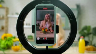 Aşçılık programının kaydı. Yemek blogcusu kadın mutfakta kahvaltı hazırlıyor, konuşuyor ve gülümsüyor, akıllı telefondan akıllı telefon ile yemek videosu kaydediyor..