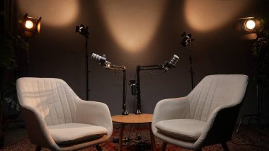 Düşünceli bir podcast stüdyosunda, iki şık sandalye birbirine dönük bir masada, mikrofonlar meşgul edici bir röportaj için hazır. Yumuşak ışık davet edilen alanı vurgular.